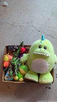 Top-down image showing a box of assorted stuffed animal toys including large green monster plush, brightly colored bird, and other multi-colored animals