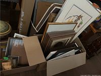 Multiple framed and unframed prints and artworks stored stacked inside a cardboard box and leaning against furniture.