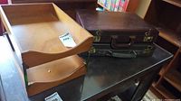 Two briefcases and two wooden office trays placed on a black wooden table. Briefcases have brass locks and handles. Trays have a smooth finish and curved edges for paper organization.
