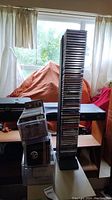 Photo showing tall black CD tower filled with CDs next to several smaller stacks of CDs in jewel cases on a table near a window.