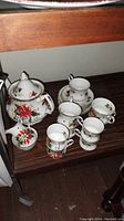 Photo showing teapot with lid, cream pitcher, sugar bowl, and four teacups with saucers adorned with Christmas floral patterns in red, green, and gold.