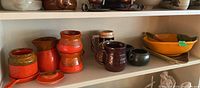 Photo showing ceramic set on shelf with stackable mugs, sugar bowl with lid, creamer, candlestick, beer stein, small black pottery piece, and two serving dishes