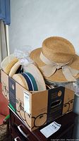 Box containing eight straw hats stacked, showing variety of ribbon colors and styles.