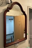 Wall mounted mirror with wood frame and arched top design showing reflection of interior space including door and furniture.