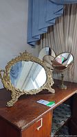 Three tabletop mirrors on wooden desk, two round tilt mirrors and one large ornate gold framed oval mirror.