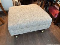 Front and top angled view of a square ottoman upholstered in beige fabric with wooden legs on brass caster wheels, showing overall shape and fabric texture.
