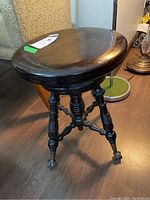 Front perspective of the wooden swivel piano stool showing the circular seat and ornate turned legs ending in metal claw feet with glass caster wheels.
