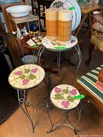 Two metal plant stands with mosaic grapevine tops and two insulated beverage containers on a small round table in a room with various furniture.