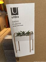 Close up photo of the Umbra Grove Planter product label on the packaging, showing a white rectangular planter elevated on wooden legs with plants inside.