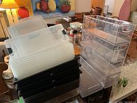 Photo showing clear plastic stacking shoebox bins with lids stacked on top of black plastic bins without lids on a table