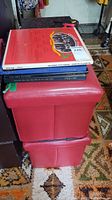 Stack of various vinyl records on top of a red upholstered storage ottoman consisting of two cubes.