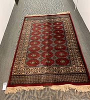 Full view of the Persian style wool carpet laid out on a carpeted floor, showing the main red field with geometric patterns and cream-colored fringe on one end.