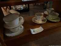 Photo of four teacups and six saucers on wooden shelf, showing various designs and gold trims.