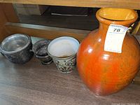 Four ceramic items: one large orange vase and three smaller plant pots including one with a glass liner, placed on a wooden floor against a shelf.