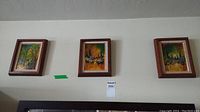 Three framed paintings on board hung on a wall, showing urban cityscapes with colorful abstract street scenes.