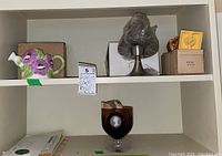 Shelf holds a ceramic watering can with floral design, a metal tealight lamp with shade, a Boston Bruins bobble head, and a small glass piece.