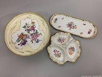 Set of three porcelain serving dishes with floral hand-painted designs and gold trim arranged together on a gray background