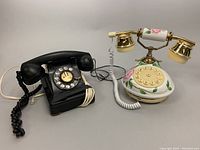 Two telephones side by side: a black Western Electric rotary phone and a white floral decorative rotary phone with gold accents
