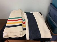 Two folded Hudson's Bay Company blankets on a black surface, one with multicolored stripes and the other with navy stripes and fringe.