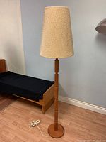 Full view of the 62-inch tall mid-century modern teak floor lamp with cone-shaped woven burlap shade on a light wooden floor near a wall and wooden furniture.