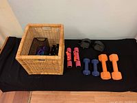 Wicker bin with athletic accessories including dumbbells, wrist weights, and pedal straps spread out on black surface.