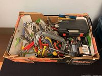 Box containing assorted hand tools including C-clamps, pliers, wire cutters, and a small black case inside a cardboard box.