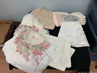 Box of assorted linens including embroidered and lace tablecloths and napkins on a surface.