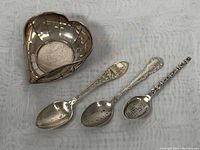 Photo of a heart-shaped silver bowl alongside three decorative sterling silver spoons showing overall lot contents.