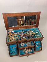 Wooden jewelry box open showing interior with blue velvet lining and assorted costume jewelry including necklaces, bracelets, earrings, and rings.