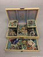Open vintage jewelry box showing compartments filled with assorted costume jewelry including necklaces, bracelets, rings and brooches.