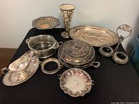 Entire lot of vintage silver plated servingware including vase, gravy boat, trays, bowls, covered serving dish, and condiment holder arranged on black surface.