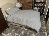 Full view of the microfiber suede full bed with bedding and pillows in a guest room setup, showcasing the bed's frame and headboard design against a brick wall and furniture in the background.