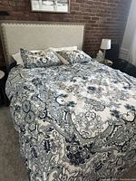 Full view of queen size bed with patterned bedding, upholstered headboard with nailhead trim, and brick wall background.