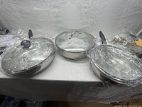 Three new stainless steel cooking pans with glass lids wrapped in plastic, shown side by side on a table.