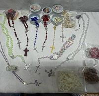 Photo of fourteen rosaries in various colors and bead materials, laid out on a white cloth, showing crucifixes and some in plastic bowls or packaging.