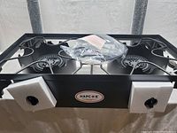 Front view of the black metal dual burner stove with two control knobs covered in foam protection, showing the burners and packaged parts on top.