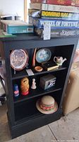Front view of black painted wooden bookcase showing three shelves with various decorative items and games on top. Paint chips visible on edges.