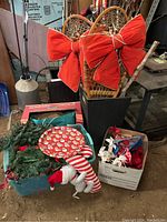 Overview of boxes, bin with Christmas decorations, red bows, greenery wreaths, and plush Santa figure near planter.