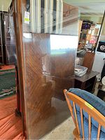Executive desk with large glass top leaning against wall. Wooden desk has chevron veneer pattern and polished finish.