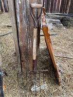 Full view of hay fork leaning against wooden beam in barn, showing wooden handle and metal tines with some bird droppings.