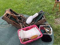 Photo showing wooden saddle riser, grooming brushes in pink basket, leather girth and reins on green outdoor surface
