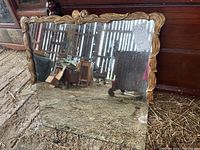 Full square mirror showing the overall wooden carved frame and reflective surface placed leaning against a wood backdrop with visible barn interior reflection.