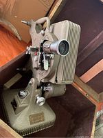 Side angle of Keystone 6mm movie projector inside carrying case showing textured gray metal body and lens.