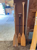 Photo showing three wooden paddles of different heights leaning against a cardboard backdrop in a garage setting.