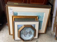 Stacked framed artworks showing a variety of frame shapes and colors, including large rectangular prints and smaller framed items