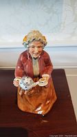 Front view of the Royal Doulton figurine showing an elderly woman holding a teapot and a teacup, seated or standing.
