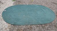 Green and cream braided oval rug displayed flat on gravel surface, showing full oval shape and size.