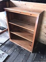 Front and side view of wooden shelving unit with three shelves placed against a wooden wall, showing overall condition and visible scratches.
