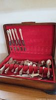 Open wood flatware box showing a variety of silver plated flatware including spoons, forks, knives, and ladles arranged in a red velvet lined interior. Visible scratches on flatware.