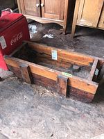 View of wooden trough placed on dirt floor next to old wooden cabinetry and Coca Cola cooler, clearly showing its rectangular shape, wood grain, and weathered condition.